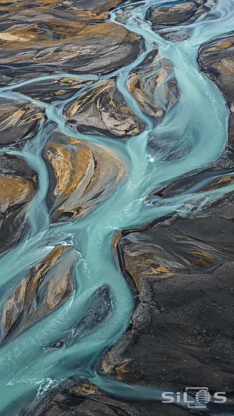 Pale turquoise channels divide across dark gravel and ochre sand in an aerial river view