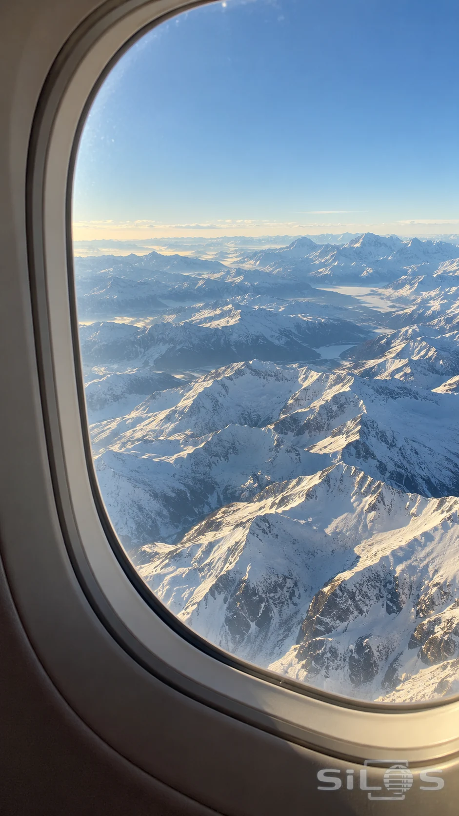 An oval airplane window frames layered snow-covered mountain ridges beneath a clear pale blue sky.