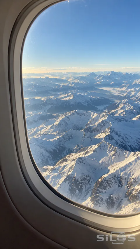 An oval airplane window frames layered snow-covered mountain ridges beneath a clear pale blue sky.