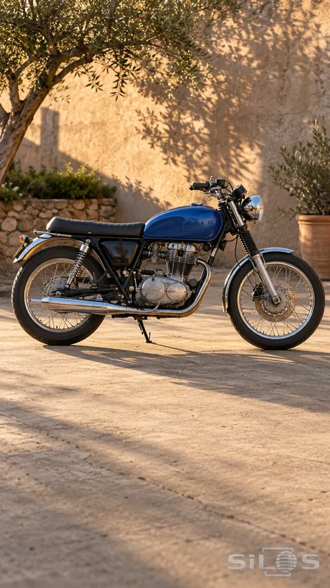 A cobalt-blue classic motorcycle with chrome parts and two spoked wheels stands in a warm sunlit stone courtyard.