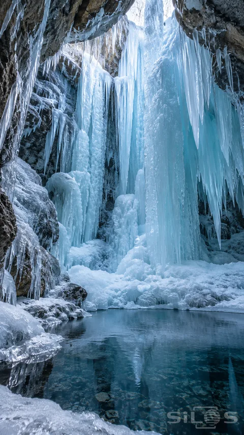 Long blue-white ice columns hang above a dark pool inside a frozen grotto