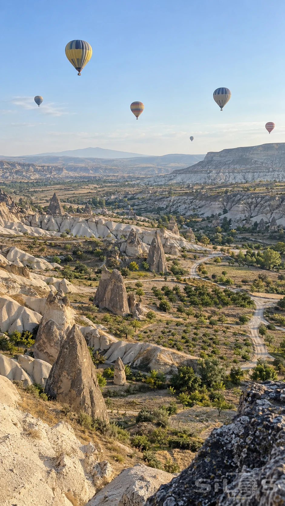 Six distant hot-air balloons drift over folded volcanic ridges, stone spires and small orchard plots.
