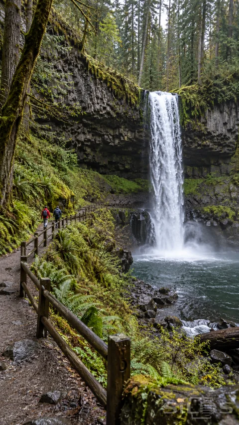 Two small hikers walk on a railed forest trail beside a waterfall dropping over basalt into a dark pool.
