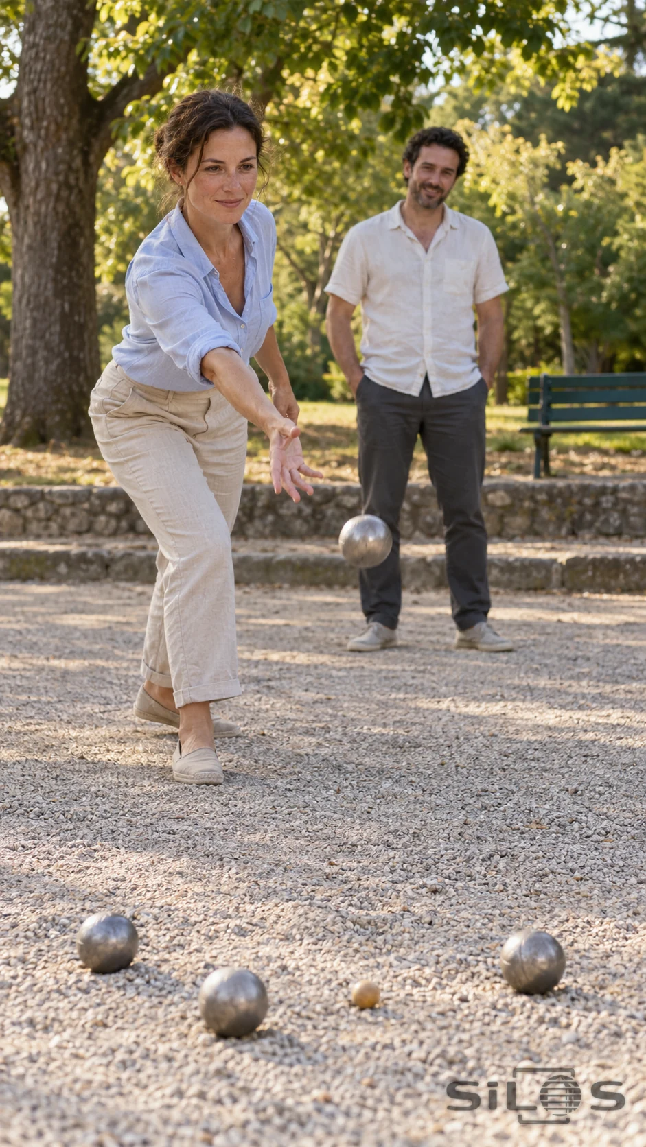 Two adult friends share a relaxed game of boules on a gravel court beneath leafy trees.