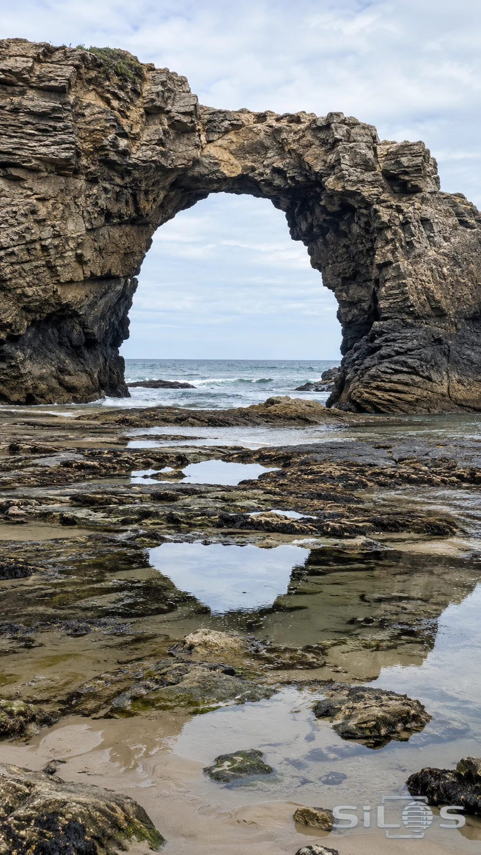 A broad coastal rock arch stands above shallow low-tide pools and a quiet sea horizon.
