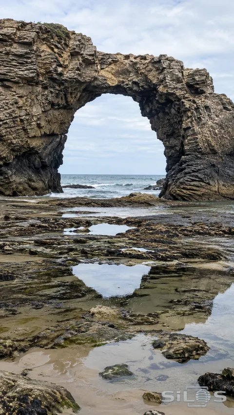 A broad coastal rock arch stands above shallow low-tide pools and a quiet sea horizon.