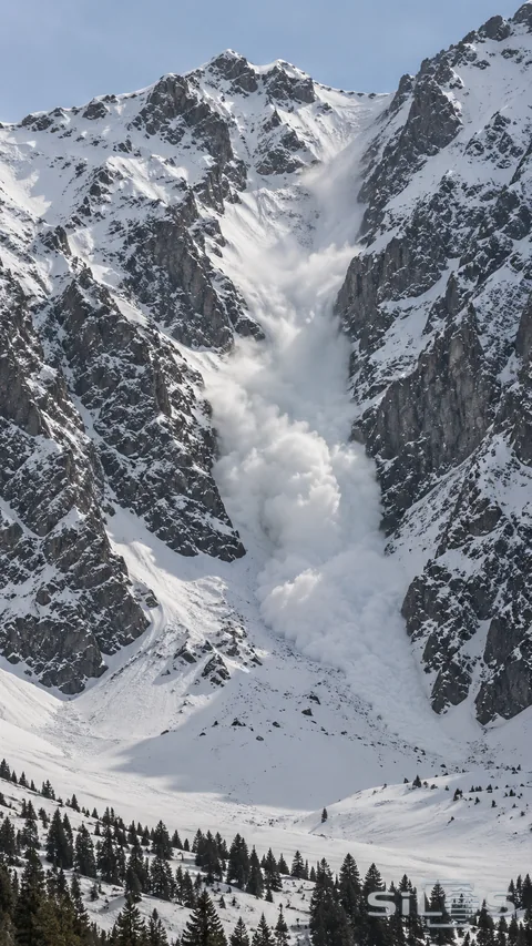 A powder-snow avalanche descends a distant empty gully between steep snowy mountain walls.