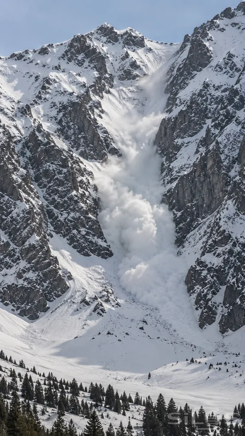 A powder-snow avalanche descends a distant empty gully between steep snowy mountain walls.