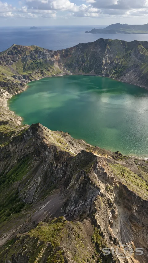 An aerial view reveals an emerald crater lake enclosed by rugged volcanic ridges.