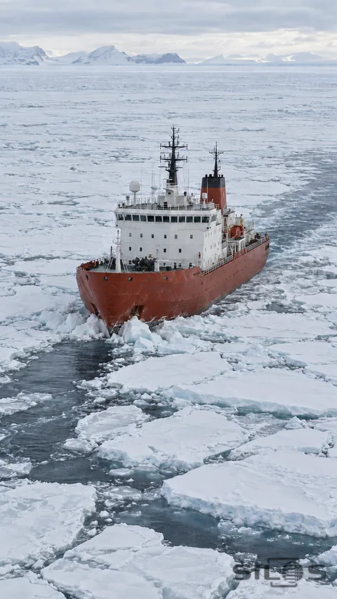 An unbranded icebreaker advances through pale fractured sea ice, leaving a narrow dark channel behind.