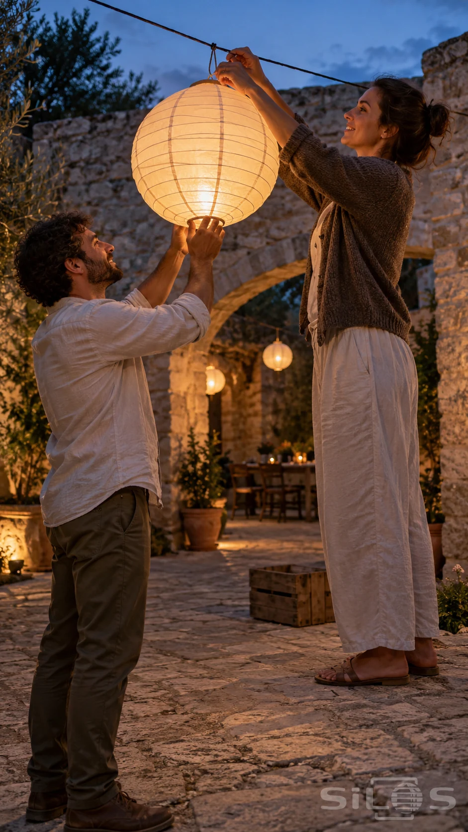 Two adults hang a handmade lantern as a quiet courtyard gathers evening light.