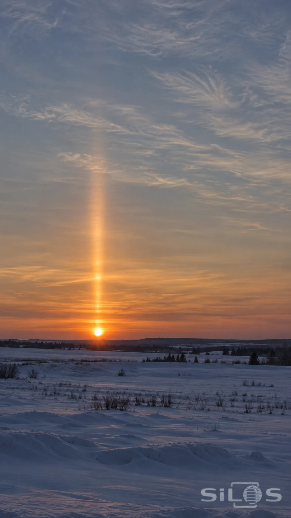 A narrow softly edged sun pillar rises above the low sunset over a quiet snowy plain.