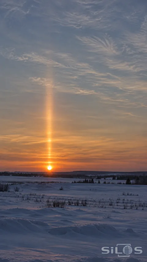 A narrow softly edged sun pillar rises above the low sunset over a quiet snowy plain.