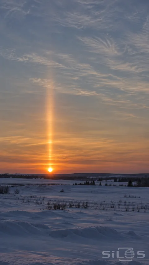 A narrow softly edged sun pillar rises above the low sunset over a quiet snowy plain.