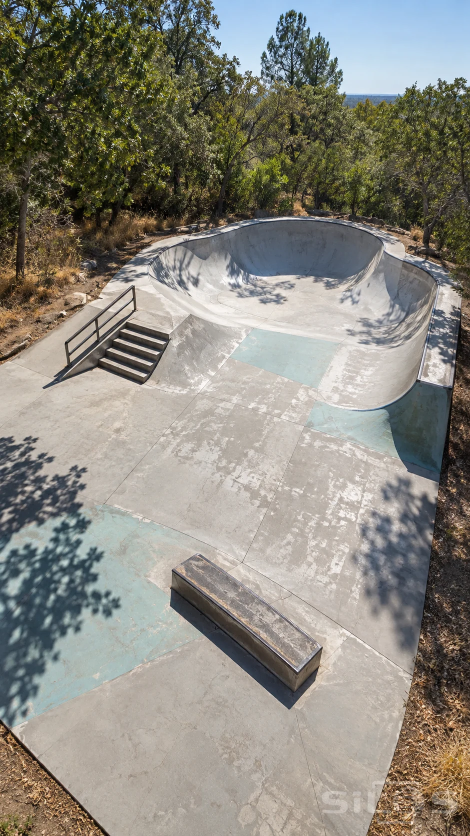 An empty worn concrete skatepark has a broad curved bowl, a short stair set with rail and a low grind box among leafy trees in daylight.