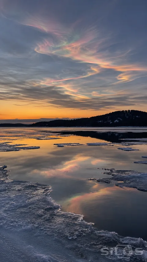 Subtle pink, green and gold iridescent cloud bands cross a twilight sky above partly frozen water, with an amber horizon and dark wooded shore.
