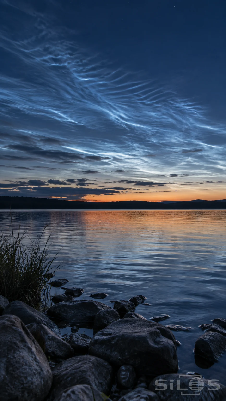Thin blue-white cloud ripples above a dark lake at twilight, with an amber horizon, distant wooded ridge and wet foreground stones beside reeds.