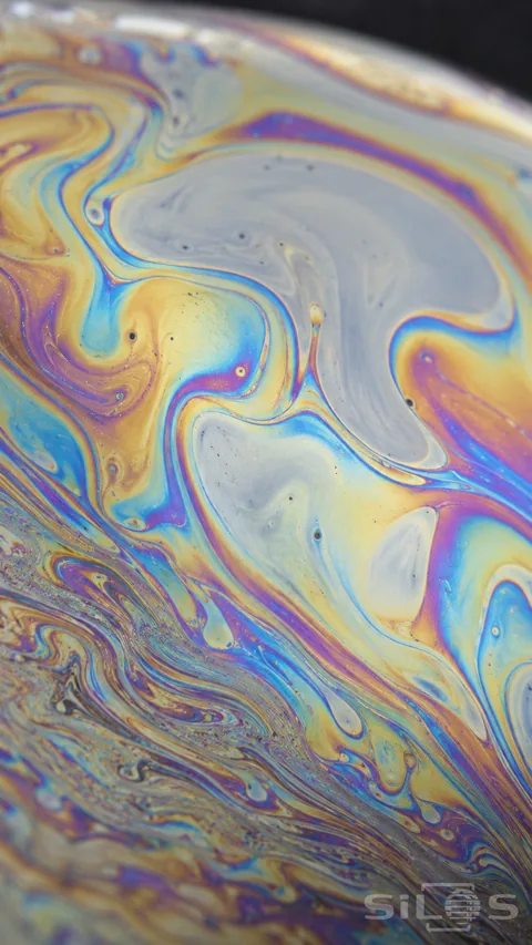 Fine blue; yellow and purple interference fringes curl across a thin soap film; with pale translucent regions; tiny irregular spots and a softly receding curved margin.