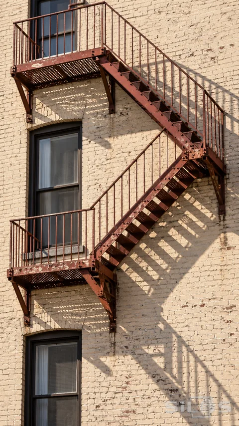 A red steel fire escape casts angular afternoon shadows across a pale brick wall