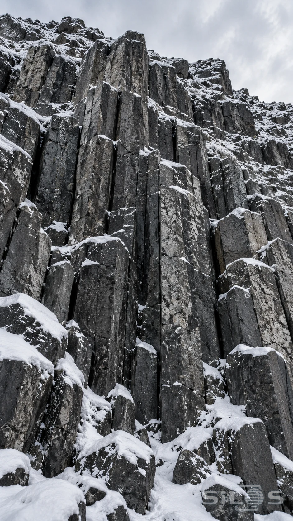 Fresh snow settles into the ledges of dark basalt columns beneath an overcast sky