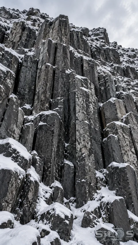 Fresh snow settles into the ledges of dark basalt columns beneath an overcast sky