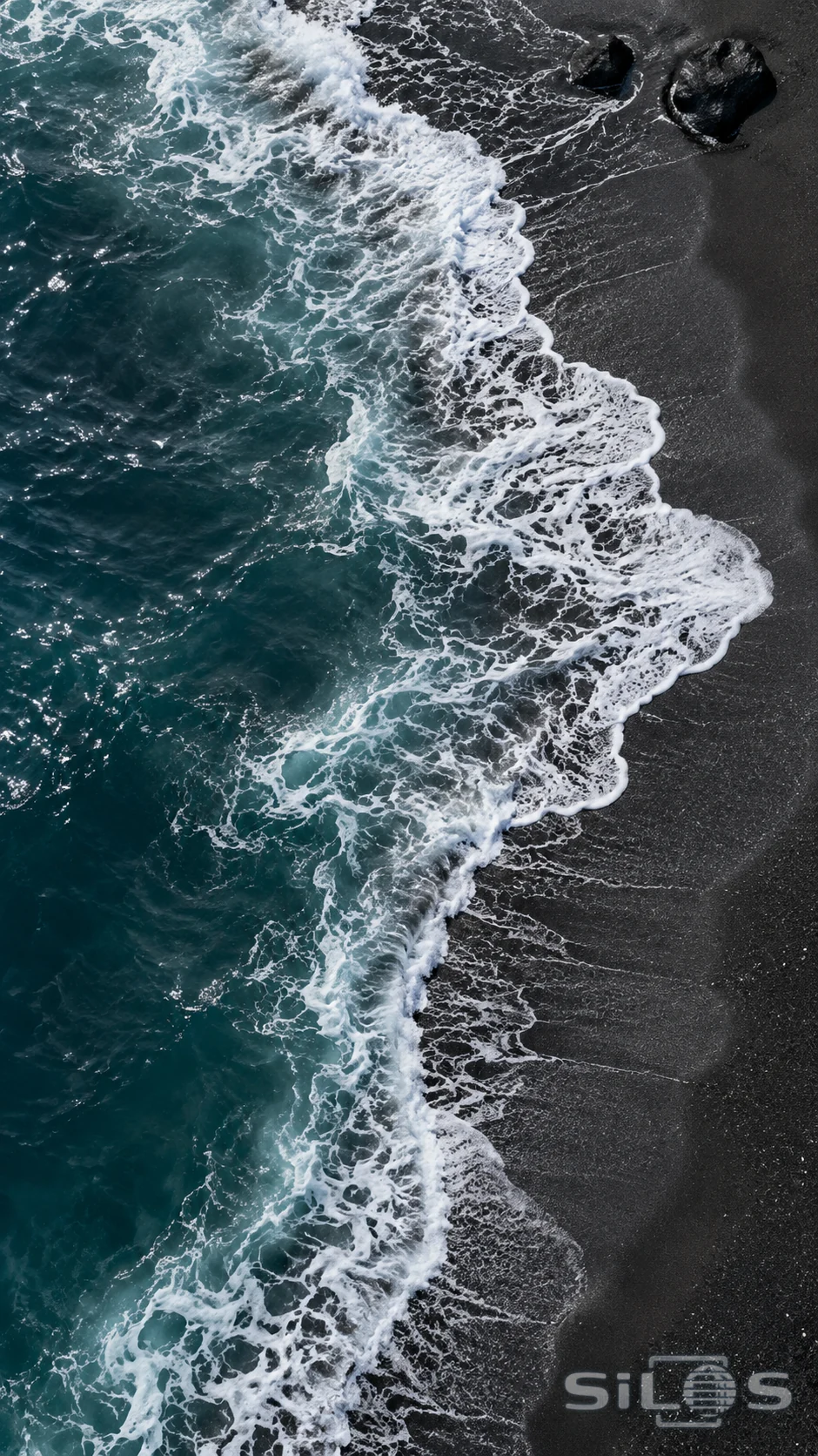 A curved lace of white surf traces a black sand shoreline beside deep teal water