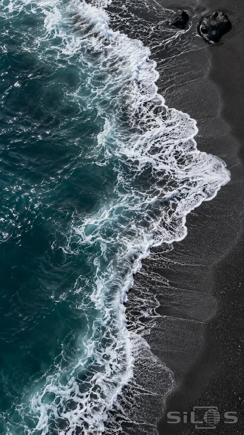 A curved lace of white surf traces a black sand shoreline beside deep teal water