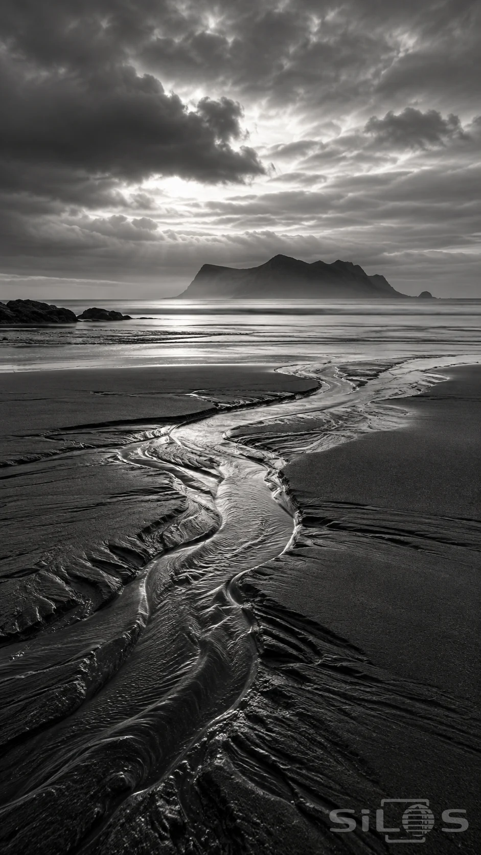 A winding tidal channel crosses dark wet sand toward a misty island and a bright opening in coastal cloud.