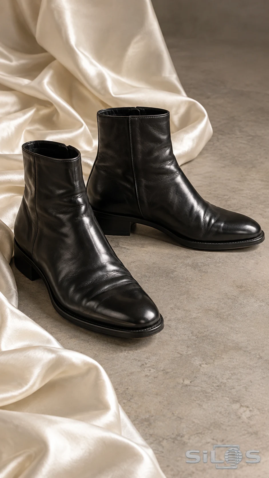 A pair of black leather ankle boots stands beside flowing ivory satin on a gray floor.
