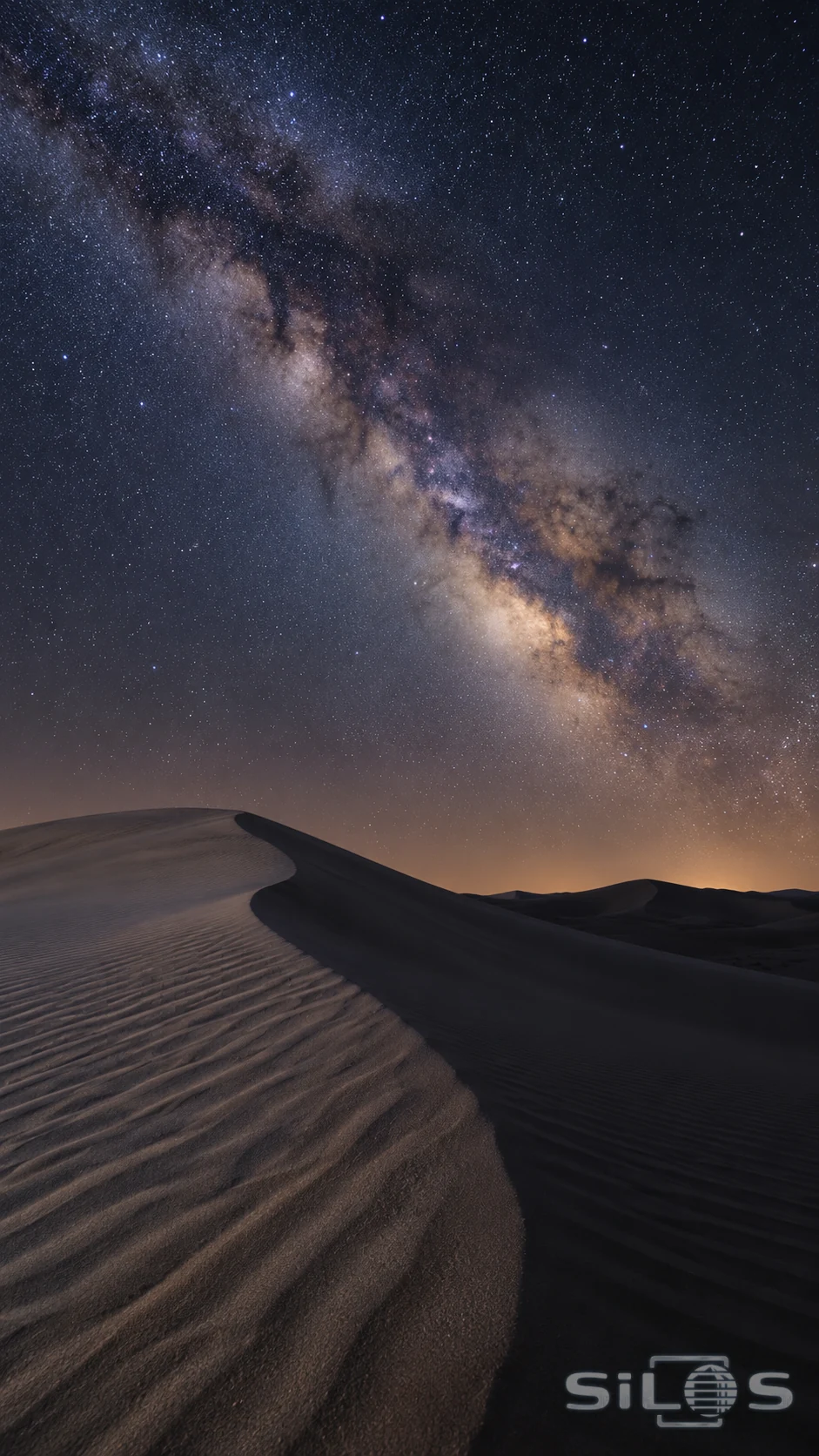 The Milky Way and small stars fill a blue-black night sky above curved sand dunes and sharply textured ripples.