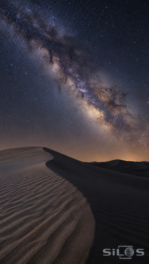 The Milky Way and small stars fill a blue-black night sky above curved sand dunes and sharply textured ripples.