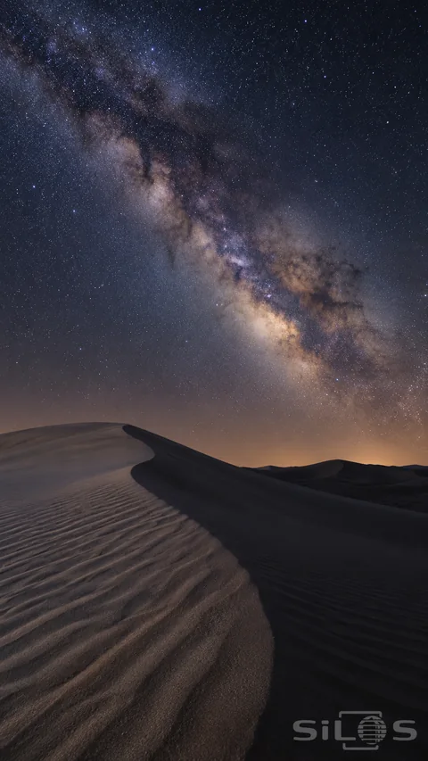 The Milky Way and small stars fill a blue-black night sky above curved sand dunes and sharply textured ripples.