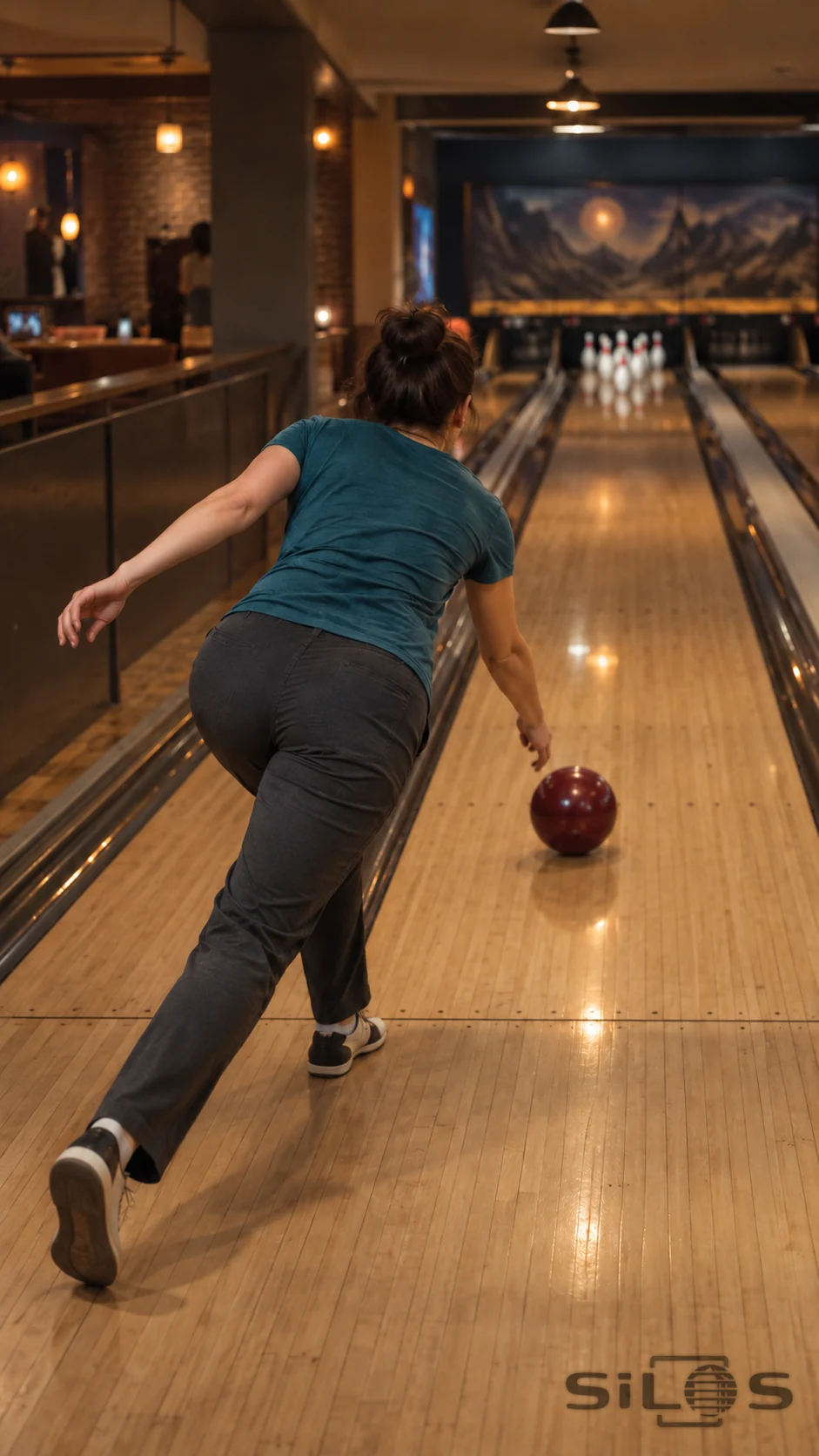 An adult woman in a muted teal shirt follows through after releasing one dark-red ball on a honey-colored wooden bowling lane, with softly focused pins at the far end.