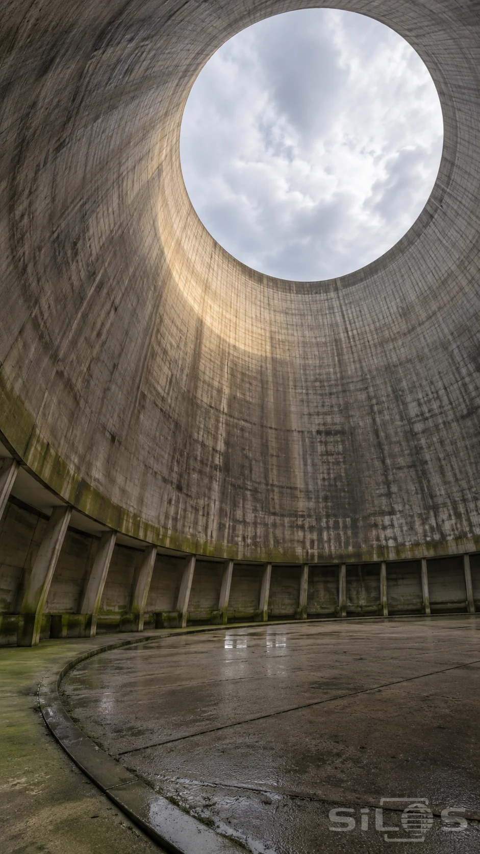 A low upward off-axis view inside an empty concrete cooling-tower shell shows its complete elliptical sky opening, continuous curved wall, connected supports and damp circular floor.