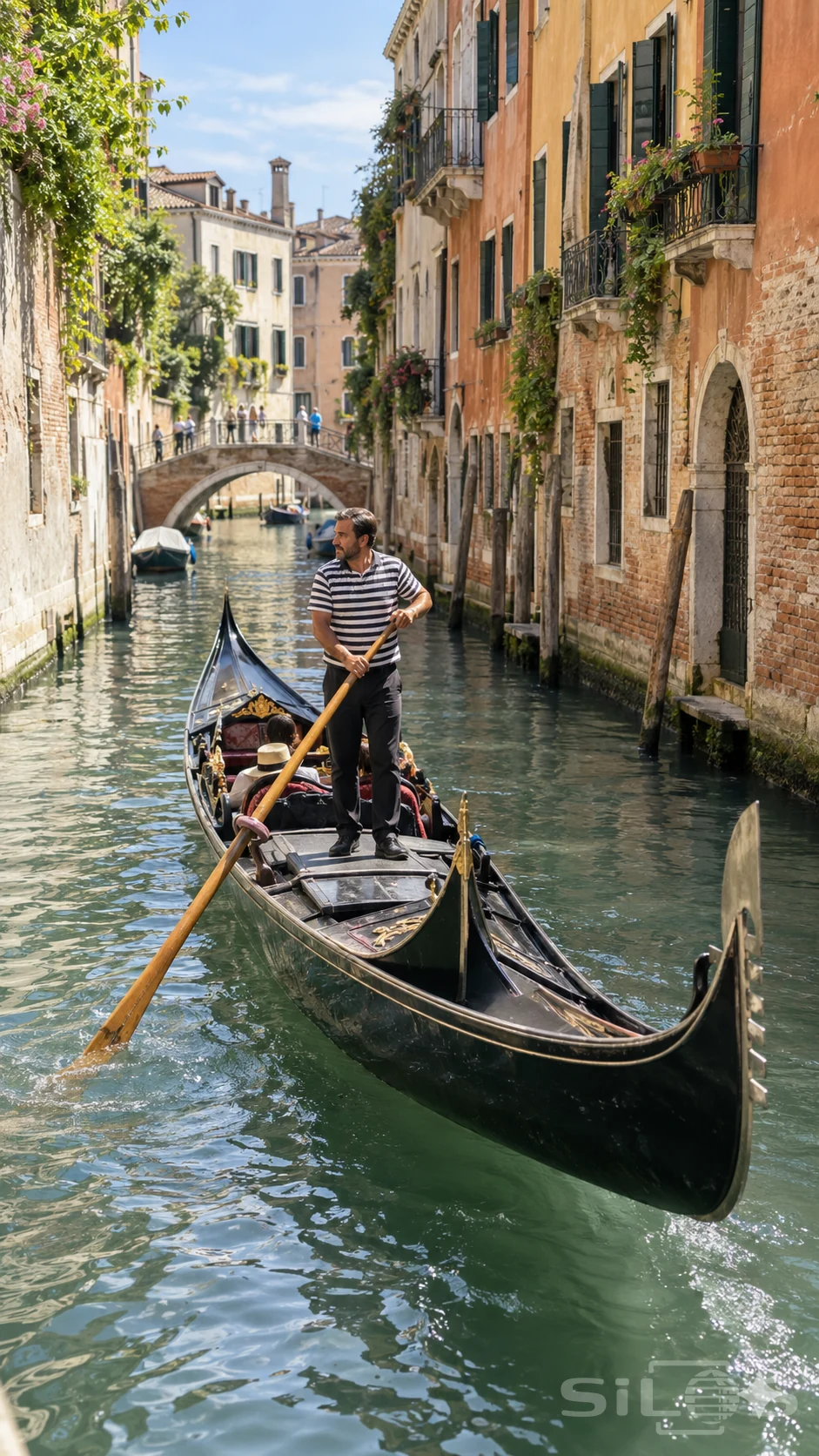 A gondolier guides a black boat along a sunlit canal between weathered buildings and an arched footbridge.