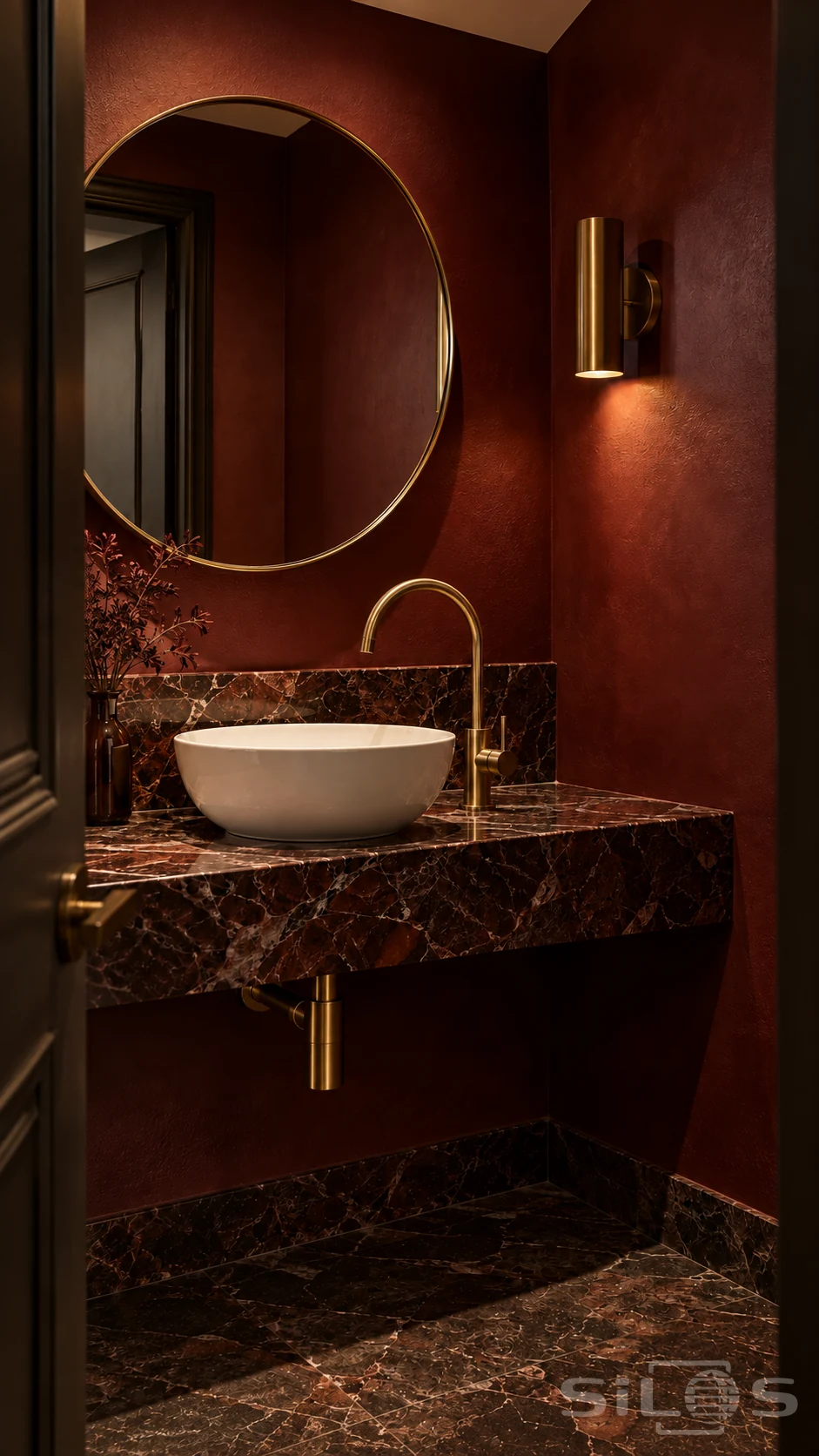 Warm brass light illuminates a round mirror, arched faucet and porcelain basin above a red-marble vanity in a deep oxblood powder room.