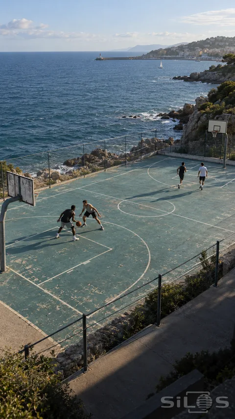 Four young adults play basketball on a weathered coastal court above rocky water, with a seaside town and breakwater in the distance.