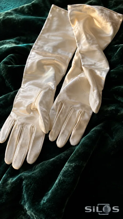 A pair of long ivory silk evening gloves arranged diagonally across folded dark green velvet.