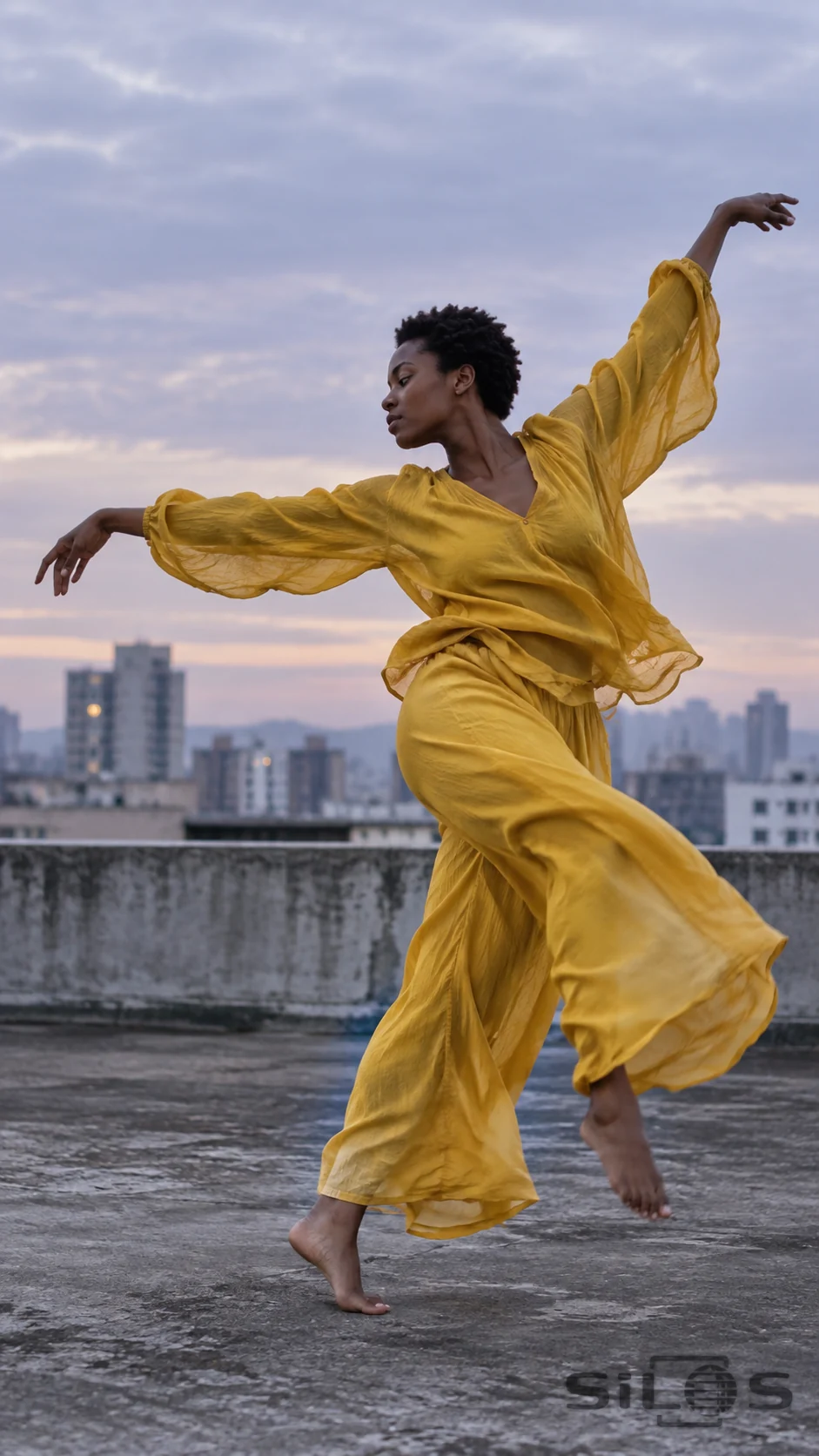 Adult dancer in loose mustard clothes turning barefoot on a concrete rooftop with a soft city skyline behind.