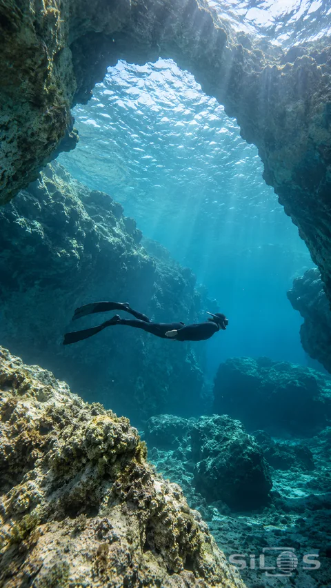 A freediver in a black wetsuit glides beneath a submerged stone arch while sunlight filters through clear blue water.