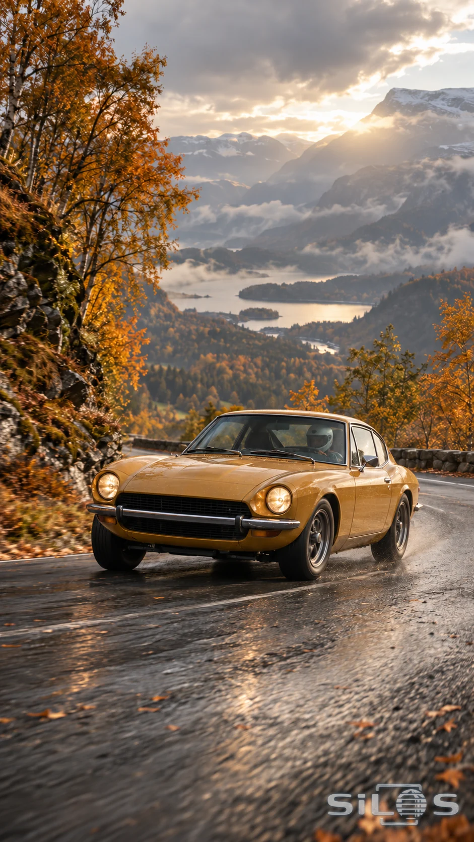 An ochre coupe follows a wet mountain hairpin below golden trees and a sunlit misty valley.