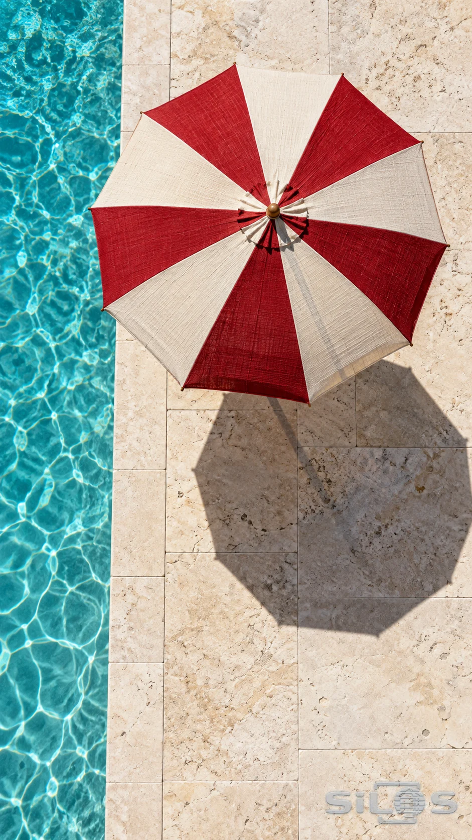 An overhead view of a red-and-white parasol and its shadow on pale stone beside turquoise water.