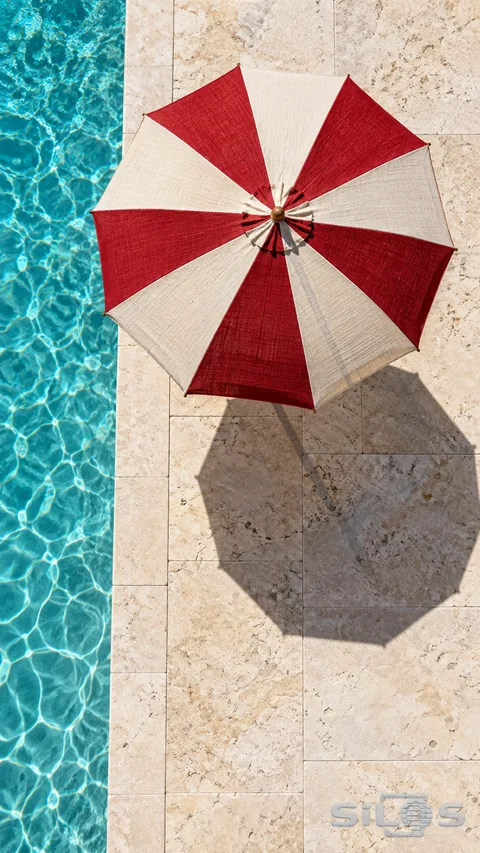 An overhead view of a red-and-white parasol and its shadow on pale stone beside turquoise water.