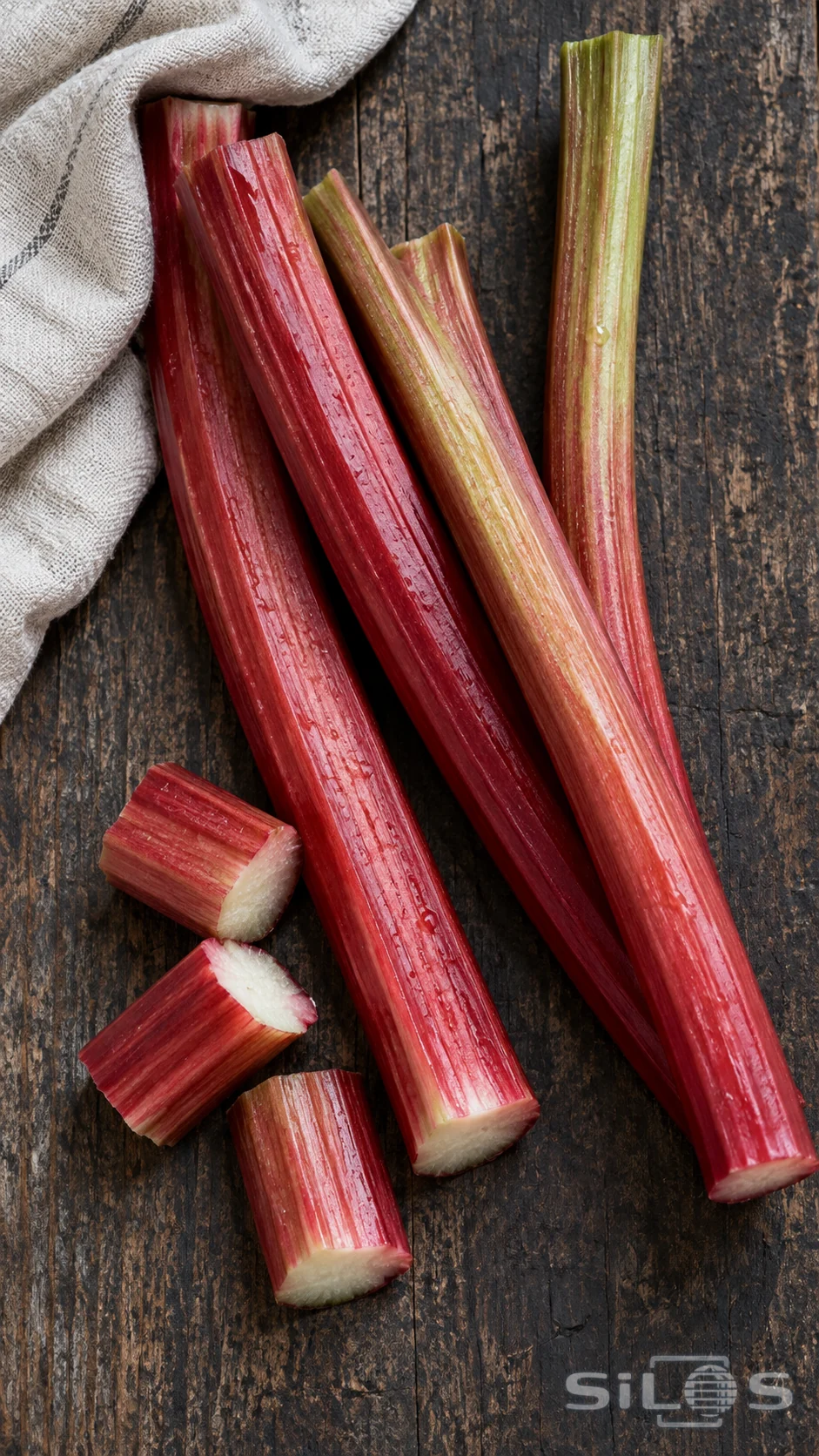 Red and green rhubarb stalks with pale cut ends rest on a dark wooden board beside linen.