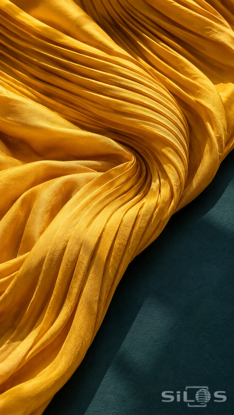 Golden saffron silk gathers into curved pleats on a dark teal tabletop in bright side light.