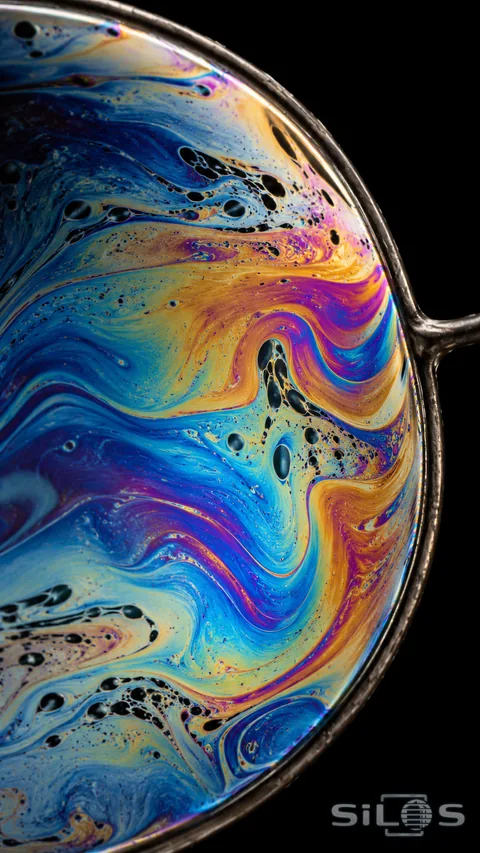 A thin soap film stretches across a metal loop against black. Cobalt, cyan, gold, and magenta flow around tiny dark cells and curved interference bands in a tightly framed macro study.